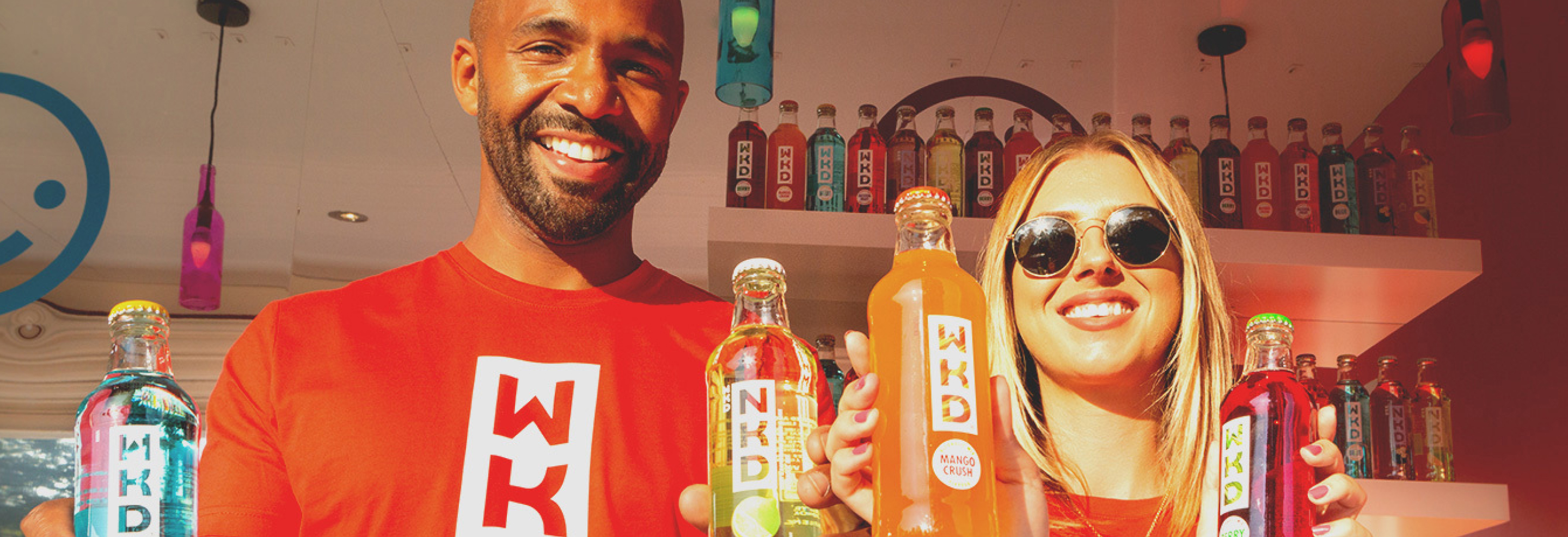 Two smiling people wearing red WKD shirts hold colourful WKD drinks in front of a display filled with bottles.
