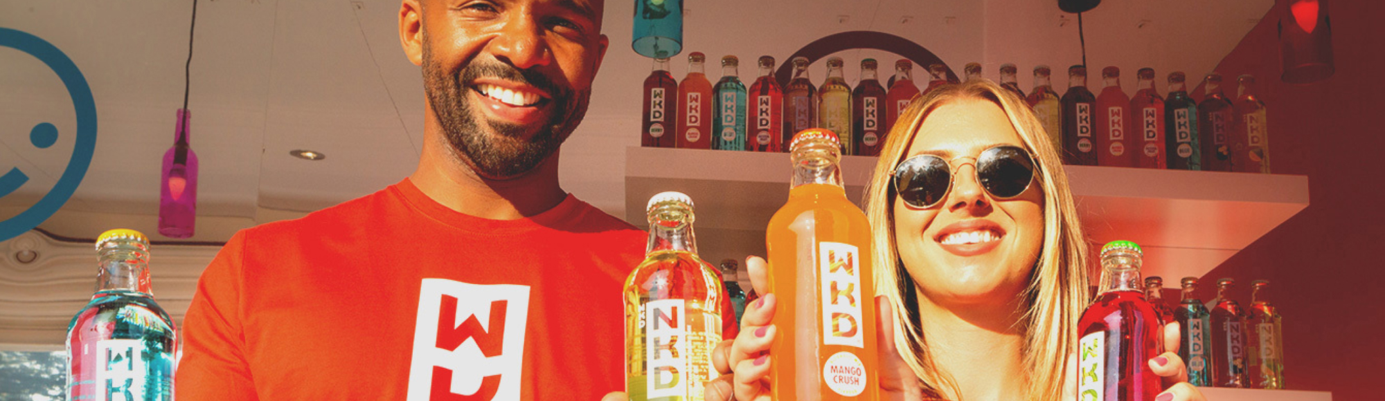 Two smiling people wearing red WKD shirts hold colourful WKD drinks in front of a display filled with bottles.