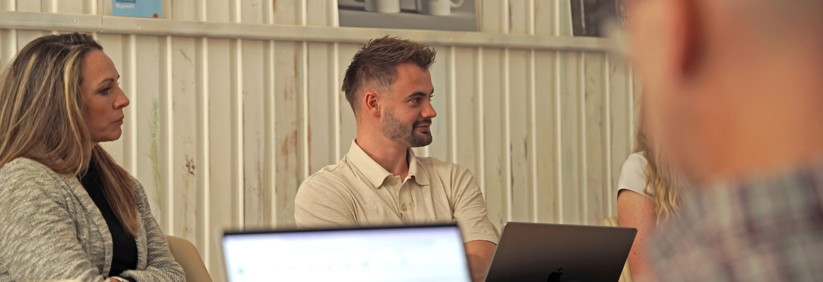 People seated around a table in a casual office setting, listening attentively with open laptops in front of them.