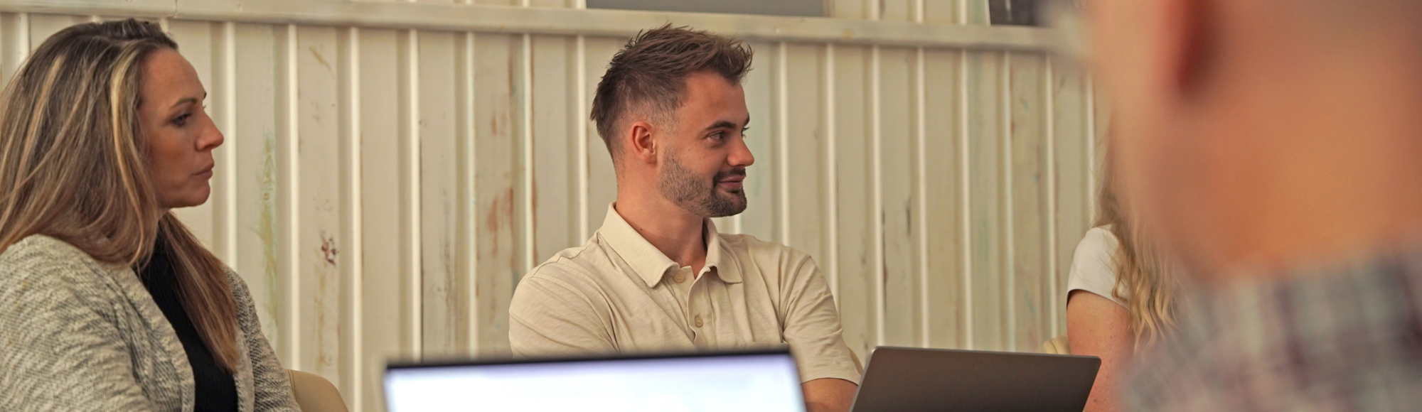 People seated around a table in a casual office setting, listening attentively with open laptops in front of them.