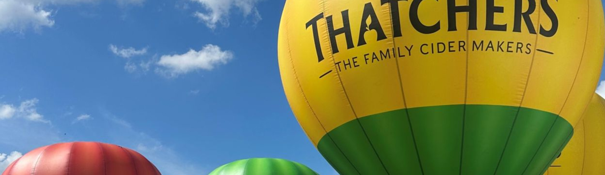 Hot air balloons in red, green, and yellow, with the largest balloon branded “Thatchers – The Family Cider Makers.”