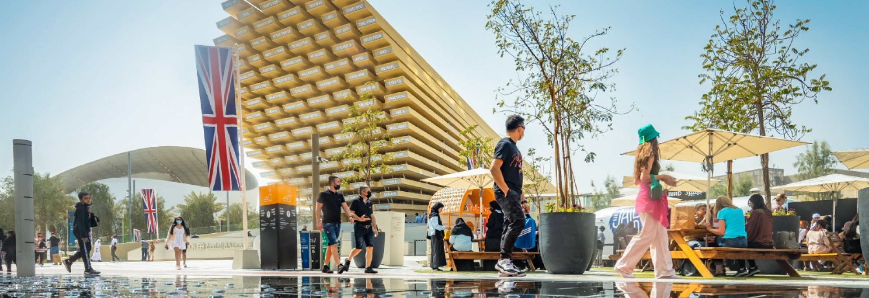 People walk and relax near the UK Pavilion at Expo, with British flags, trees, and a modern wooden structure in the background.