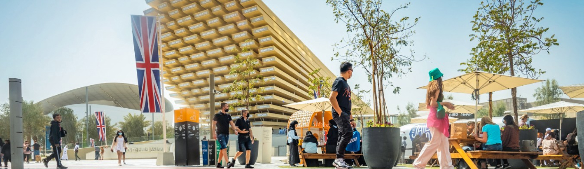 People walk and relax near the UK Pavilion at Expo, with British flags, trees, and a modern wooden structure in the background.
