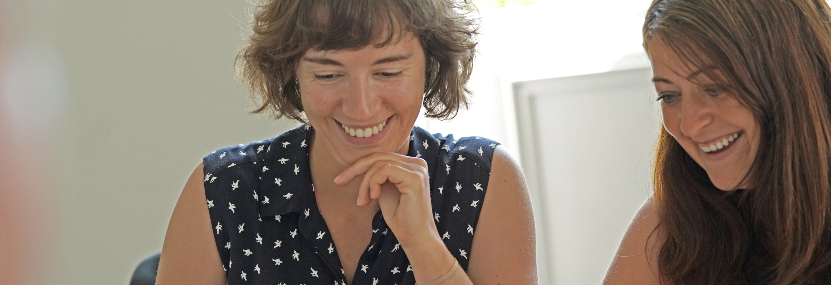 Two women smiling and looking down at something out of frame, seated in a brightly lit room.