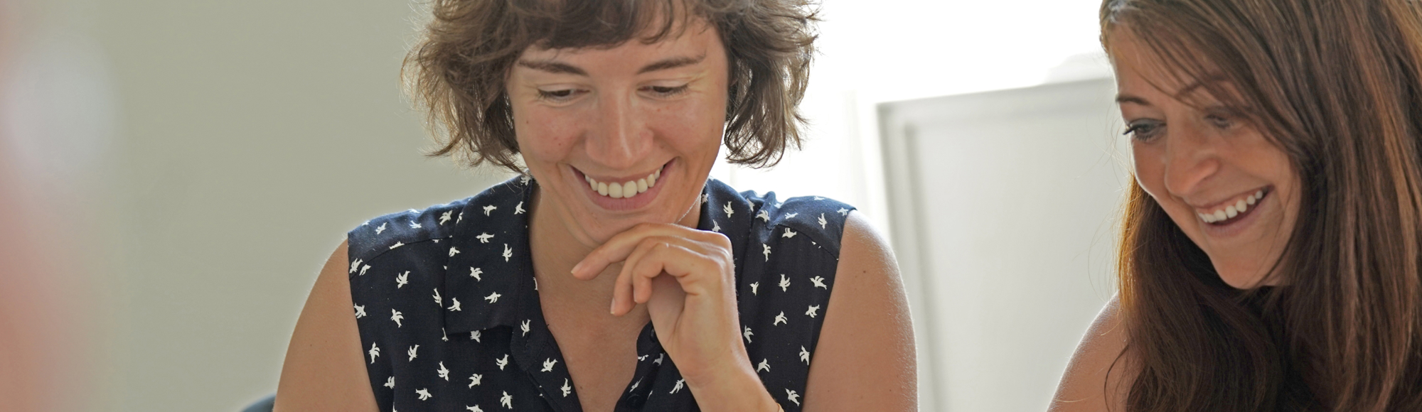 Two women smiling and looking down at something out of frame, seated in a brightly lit room.