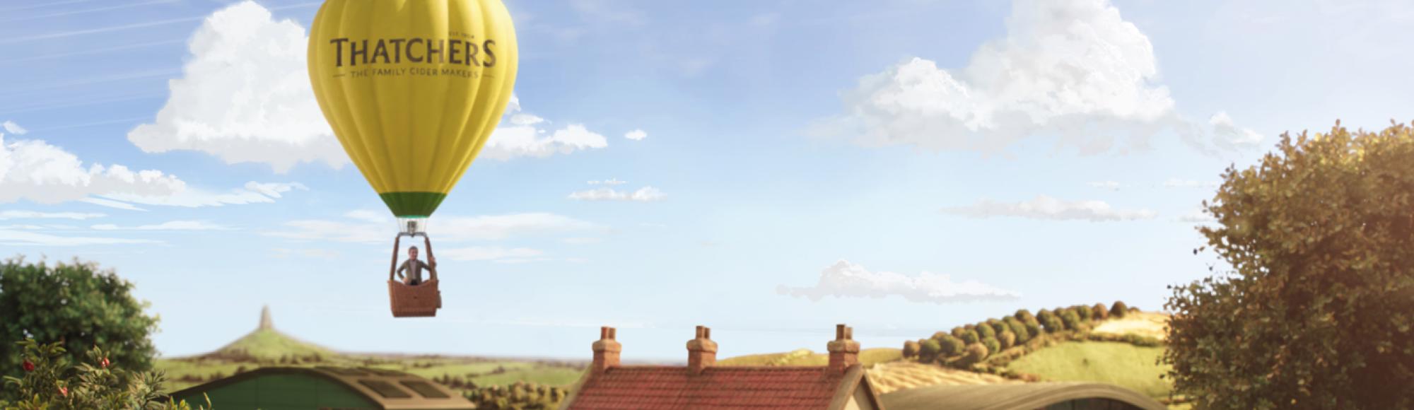 A yellow Thatchers hot air balloon floats above a countryside village with rolling hills, trees, and a farmhouse below.