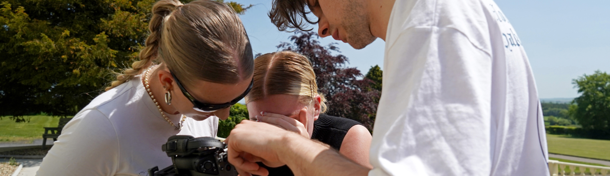 Three people closely examining a camera setup outdoors on a sunny day, with trees and a lawn in the background.
