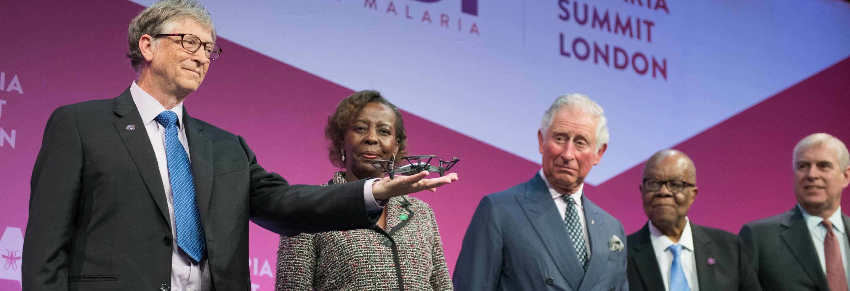 Leaders stand on stage at the Malaria Summit in London, with one person holding a small drone during the presentation.