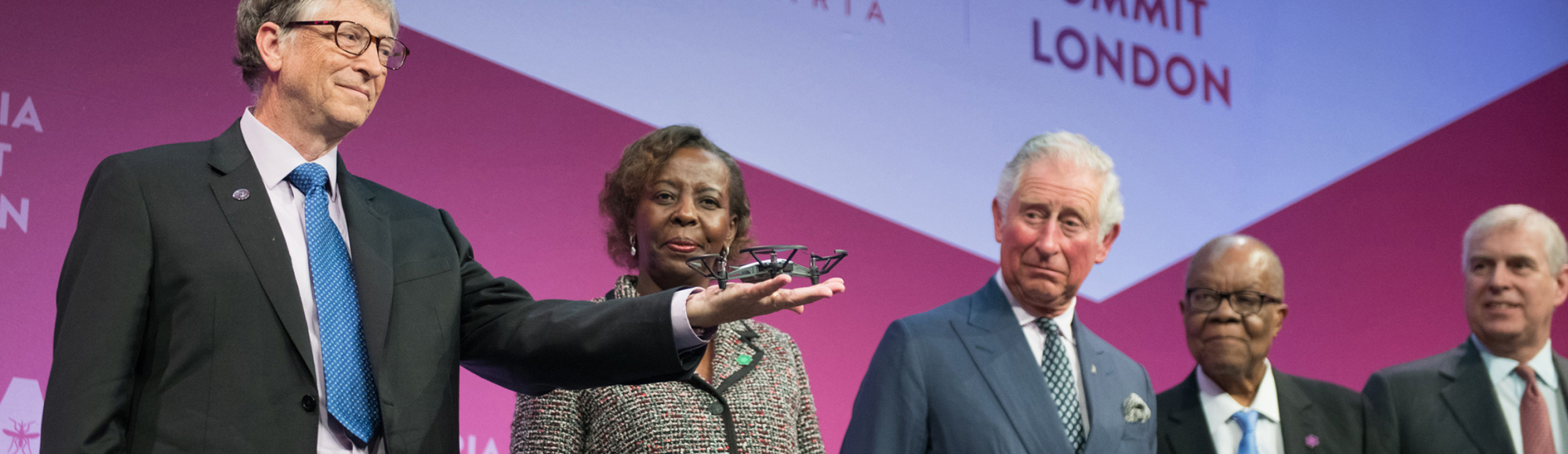 Leaders stand on stage at the Malaria Summit in London, with one person holding a small drone during the presentation.