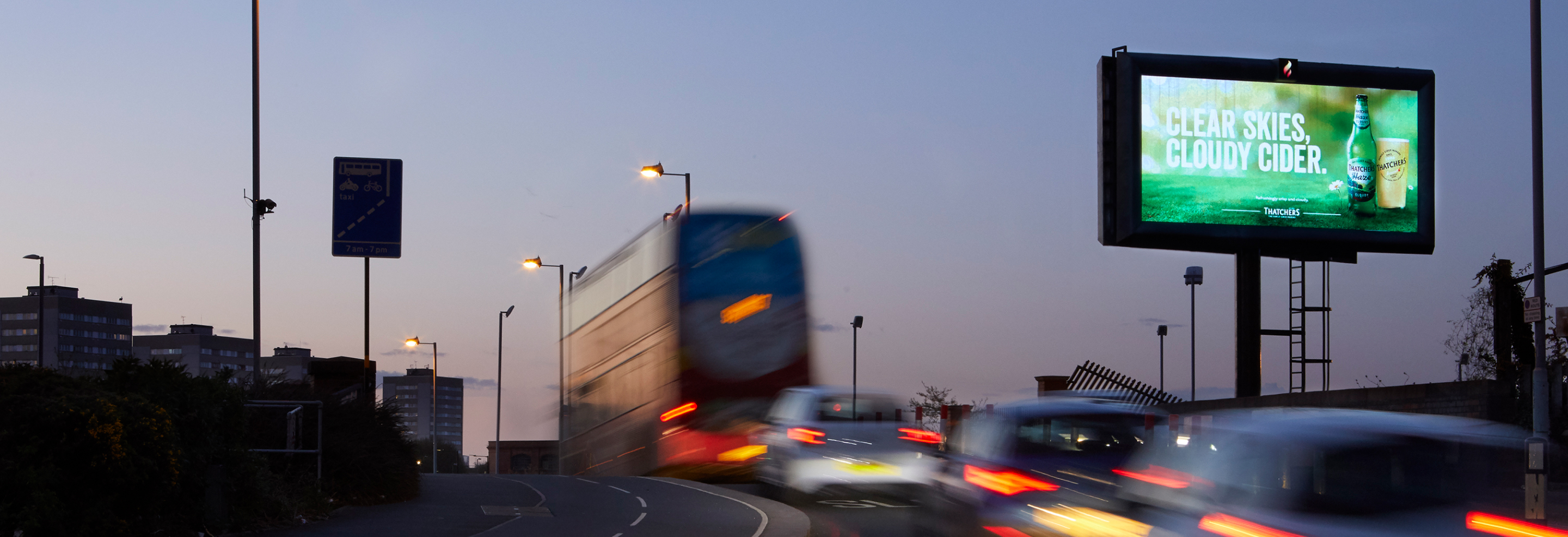 Cars and a bus pass a roadside billboard at dusk showing a cider advert that reads “Clear Skies. Cloudy Cider.”